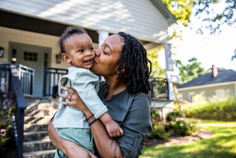 Mom Kissing Toddler By House